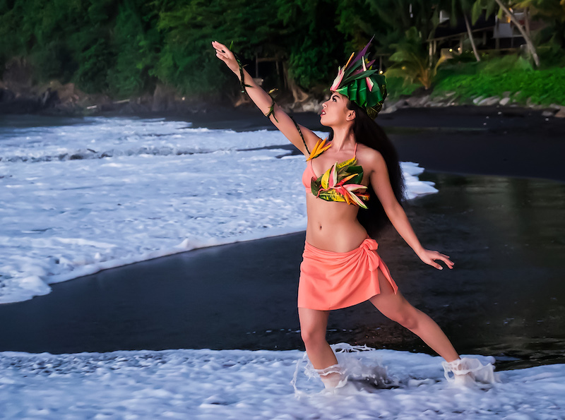 a female dancer in coral poses in the tide on a black sand beach